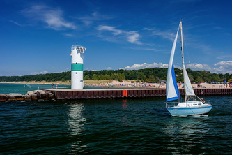 sailboat with full sail going past lighthouse on pier, beach in background
